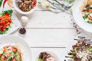 Warm salads frame on white wood flat lay. Top view on restaurant table with assortment of meat side dishes, free space. Buffet, banquet, menu concept