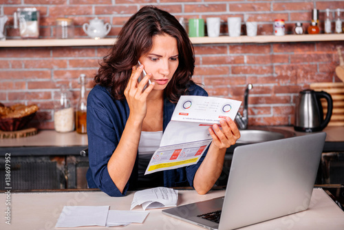 Fotografia, Obraz Young woman sitting in the kitchen holding utility bills