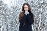 Fototapeta Koty - beautiful young woman with long dark hair sad lonely walk in the winter woods in a black jacket and mittens