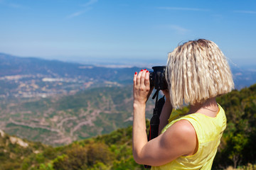 Wall Mural - Female traveler enjoying the views from the mountains of Montserrat in Spain and makes a photo on her camera. The girl in a yellow dress on background of the nature