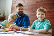 © pressmaster - Waist-up portrait of family having fun together: two funny little brothers sitting at wooden table and coloring pictures while their dad savoring the moment