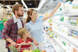 Waist-up portrait of young family with child choosing dairy products in supermarket: pretty mother stretching hand towards milk bottle while her husband and son holding shopping cart