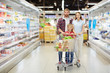 © pressmaster - Full-length portrait of confident young family with little son standing in supermarket and holding full shopping cart