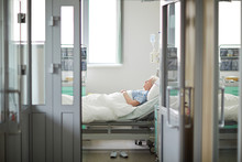 Wide Shot Image Of Doors Opened To Sterile Light Hospital Ward With Sad Lonely Old Woman Lying On Bed In The Room