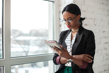 African american woman in glasses standing near the window and holding the tablet.