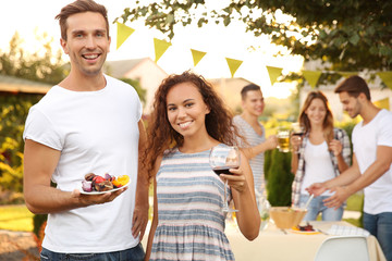 Young man and woman holding glass of wine and plate with grilled meat and vegetables outdoors