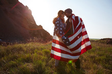 Romantic Adventure Of Young Couple Standing On Top Of Hill At Sunset, Wrapped In American Banner And Looking At Each Other Enjoying Mountain Hike
