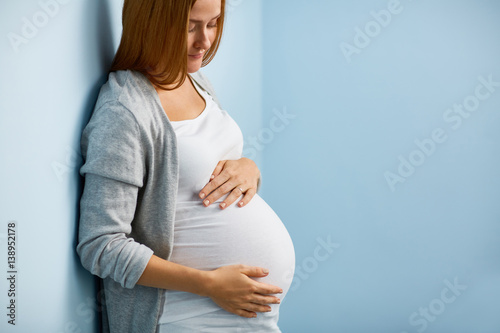 Fotografía Blond pregnant woman leaning on to blue wall caressing her big belly gently