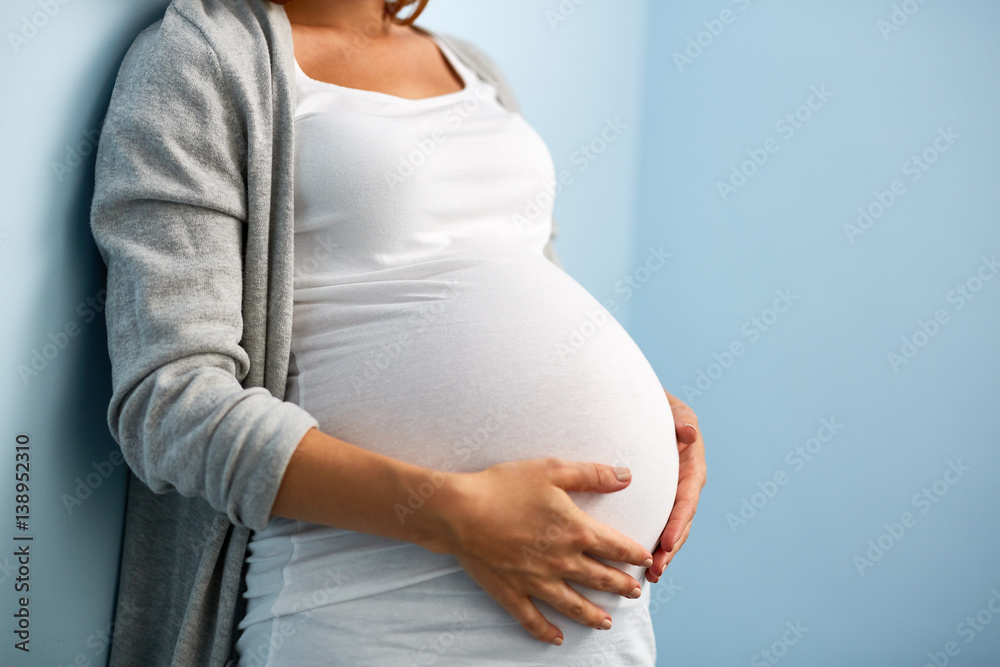 Mid-section portrait of unrecognizable woman during last months of pregnancy holding her big belly gently standing against wall in blue room - obrazy, fototapety, plakaty 