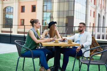 Sticker - Group of creative young business people, two pretty women and one man,  sitting at outdoor table discussing work and smiling during coffee break