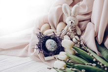 Happy Easter Greeting Card. Stylish Easter Eggs With Chick Ornaments In Nest And Bunny Rabbit And Tulips And Willow Buds On Rustic White Wooden Background. Space For Text, Soft Light
