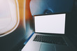 © skyNext - Wide angle shoot of modern laptop with blank screen on the blue table of airplane seat, mock up of portable computer next to porthole of flying plane with sunny day outside