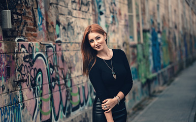 Young ginger serious looking woman posing outside dressed in dark outfit but with long statement necklace. Half-length portrait on empty street of young woman glaring away.