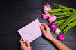 Bouquet of tender pink tulips and hands holding pink envelope on black wooden background