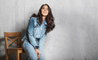 © deniskomarov - Beautiful dark haired young woman in jeans sitting and smiling on the cement wall background