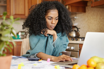 Beautiful young dark-skinned woman with Afro hairstyle wearing wrap keeping hand on touchpad of open laptop computer , looking at screen with serious concentrated expression, paying bills online