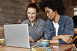 © WHstudio Leushin N - Two stylish lesbians sitting at cafe in front of open laptop, surfing internet, watching videos online during coffee break. Caucasian girl showing something on notebook pc to her African girlfriend
