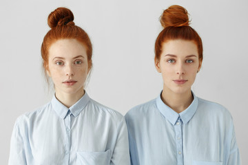 Wall Mural - Indoor shot of two gorgeous redhead girls looking alike wearing same hair buns and identical shirts having relaxed looks, smiling subtly, standing close to each other against white wall background