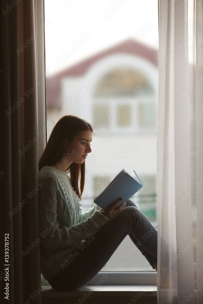 Young woman reading book on windowsill at home