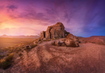 Desert with lonely rocks against multicolored cloudy sky at sunset. Blue, purple and red clouds. Panoramic. Colorful landscape with trail, stones, grass and sunlight. Nature background. Adventure