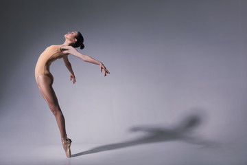 young beautiful ballet dancer in beige swimsuit posing on pointes on light grey studio background