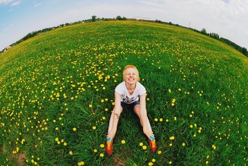 Portrait of young smiling cute blonde with short hair, he closed his eyes laughing sitting on a glade among yellow flowers of a dandelion. Beautiful landscape with the effect of fish eye .