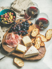 Wall Mural - Wine and snack set. Variety of cheese, olives, prosciutto, roasted baguette slices, grapes on wooden board and glasses of red wine over grey marble background, selective focus, vertical composition