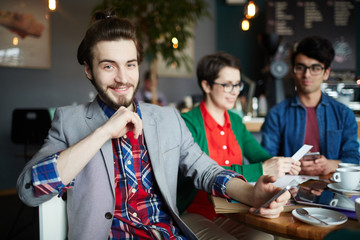 Wall Mural - Portrait of three creative people dressed in business casual at table in cafe, focus on young smiling businessman with long hair smiling cheerfully looking at camera