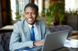 © pressmaster - Portrait of handsome African- American businessman wearing formal suit smiling cheerfully while working with laptop in cafe