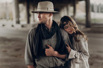 stylish hipster couple. boho gypsy woman gently hugging arm of confident man in hat under abandoned bridge. atmospheric sensual moment. rustic fashionable look.