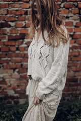 Wall Mural - Beautiful brunette hipster woman in white boho sweater and dress posing near old wall