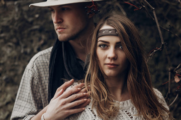 Wall Mural - Beautiful brunette gypsy girl, hipster woman in boho white sweater holding hands with handsome man in cowboy hat, free couple outdoors