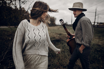 Wall Mural - stylish hipster couple posing. man in hat with guitar and his boho woman in knitted sweater in windy field. atmospheric sensual moment. fashionable look. rustic wedding concept