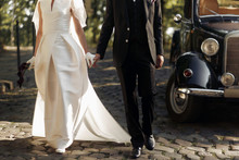 Luxury Elegant Wedding Couple Walking And Holding Hands Close Up At Stylish Black Car In Light In Park. Gorgeous Bride And Handsome Groom In Retro Style