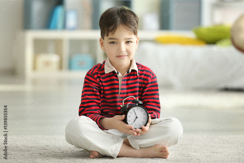 Cute little boy with alarm clock sitting on floor at home
