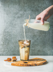 Poster - Man's hand pouring milk to Iced caramel latte coffee cocktail with frozen coffee ice cubes in glass on serving wood and marble board over grey table, dark plywood wall, selective focus, copy space