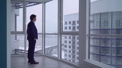 Poster - businessman in empty room with big panoramic windows looking on the urban city view. young professional man wearing in elegant suit alone in modern apartment. caucasian male deep in thought.