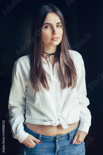 Portrait Of Pensive Sad White Caucasian Brunette Young Beautiful Girl Woman Model With Long Dark Hair And Brown Eyes In White Shirt Tied In A Knot Blue Jeans Looking In Camera On