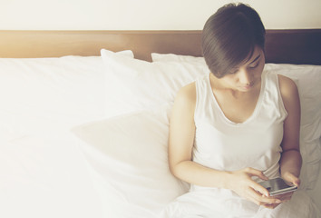 Beautiful Asian women wear white shirt sit on a bed and play mobile phone in the morning at restroom.