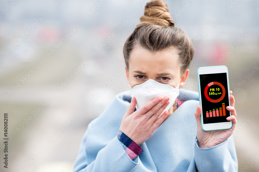 Woman in protective mask holding smart phone with air polution measurement of PM10 outdoors - obrazy, fototapety, plakaty 