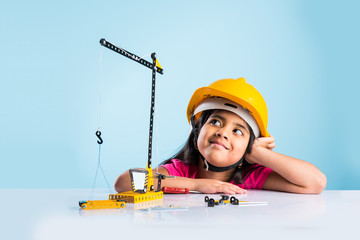 Wall Mural - cute indian baby girl playing with toy crane wearing yellow construction hat or hard hat, childhood and education concept