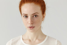 Close Up Studio Shot Of Beautiful Charming Redhead European Model With Healthy Freckled Skin Looking At Camera With Faint Smile, Posing Indoors Against Blank Wall Background, Wearing White T-shirt
