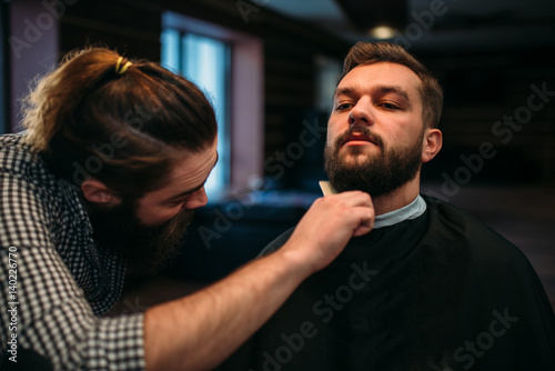 Fototapeta na szkło Barber combing beard of client man in salon cape