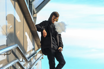 Vape. A handsome young white guy in sunglasses and in the hood is standing and smoking an electronic cigarette near the descent into the underpass on a sunny day.