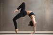 © fizkes - Young attractive yogi woman practicing yoga, standing in Bridge exercise, One legged Wheel pose, working out, wearing black sportswear, cool urban style, full length, grey studio background, side view