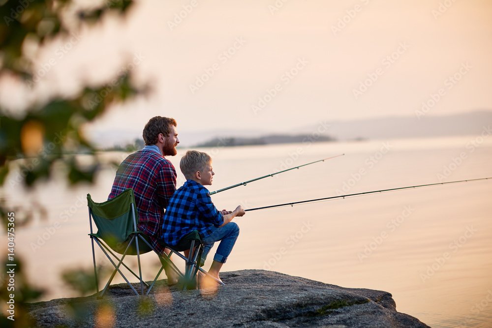 Side view portrait of father and son sitting together on rocks fishing with rods in calm lake waters with landscape of setting sun, both wearing checkered shirts, shot from behind tree - obrazy, fototapety, plakaty 