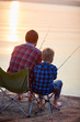 © pressmaster - Back view portrait of father and son sitting together on rocks fishing with rods in calm lake waters in sunset light, both wearing checkered shirts