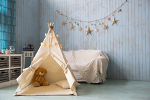 Teddy Bear Sits In A Children's House Wigwam. On The Background Of An Old Wooden Blue Wall