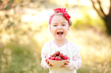 Laughing Baby Girl 4-5 Year Old Holding White Bowl With Fresh Strawberries Over Nature Green Background Outdoors. Looking At Camera. Healthy Eating. Summer Time.