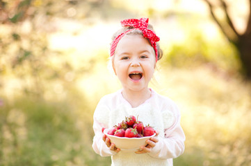 Wall Mural - Laughing baby girl 4-5 year old holding white bowl with fresh strawberries over nature green background outdoors. Looking at camera. Healthy eating. Summer time.